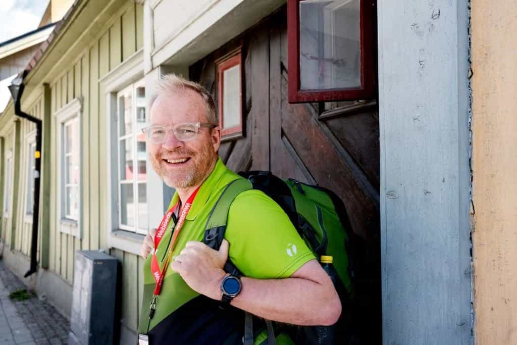 Man med ryggsäck och glasögon ler. Han står vid en trädörr