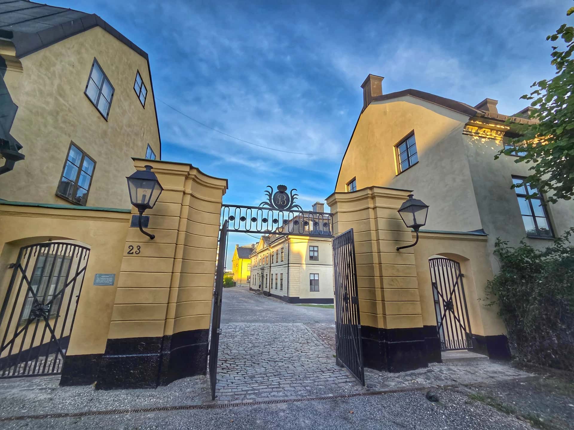 Öppen smidesjärnsgrind mellan gula byggnader. Kullerstensväg leder inåt på en stadsvandring Stockholm