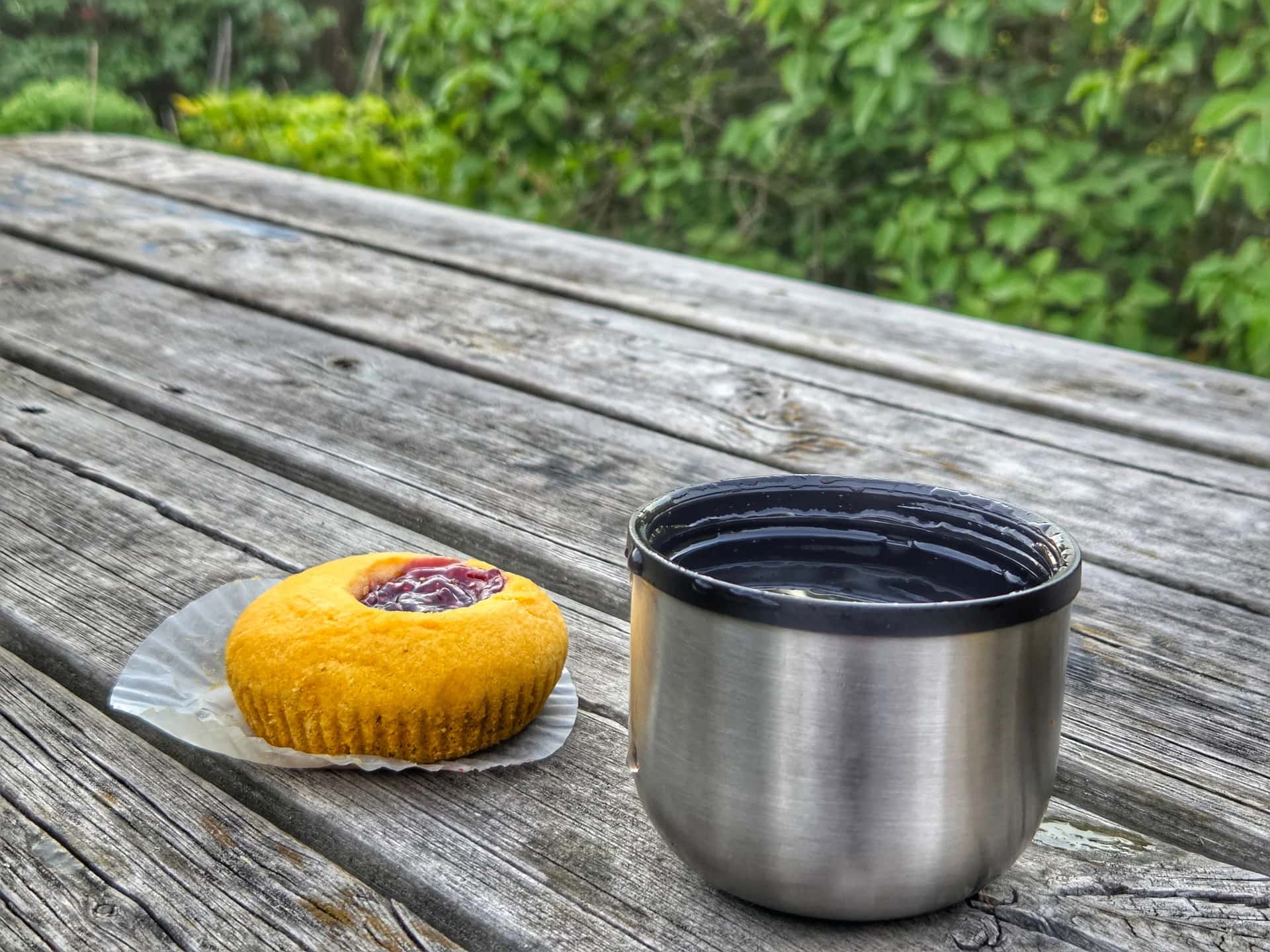 Muffin med bärfyllning och öppen termosmugg på väderbitet träbord. Grönskande bakgrund