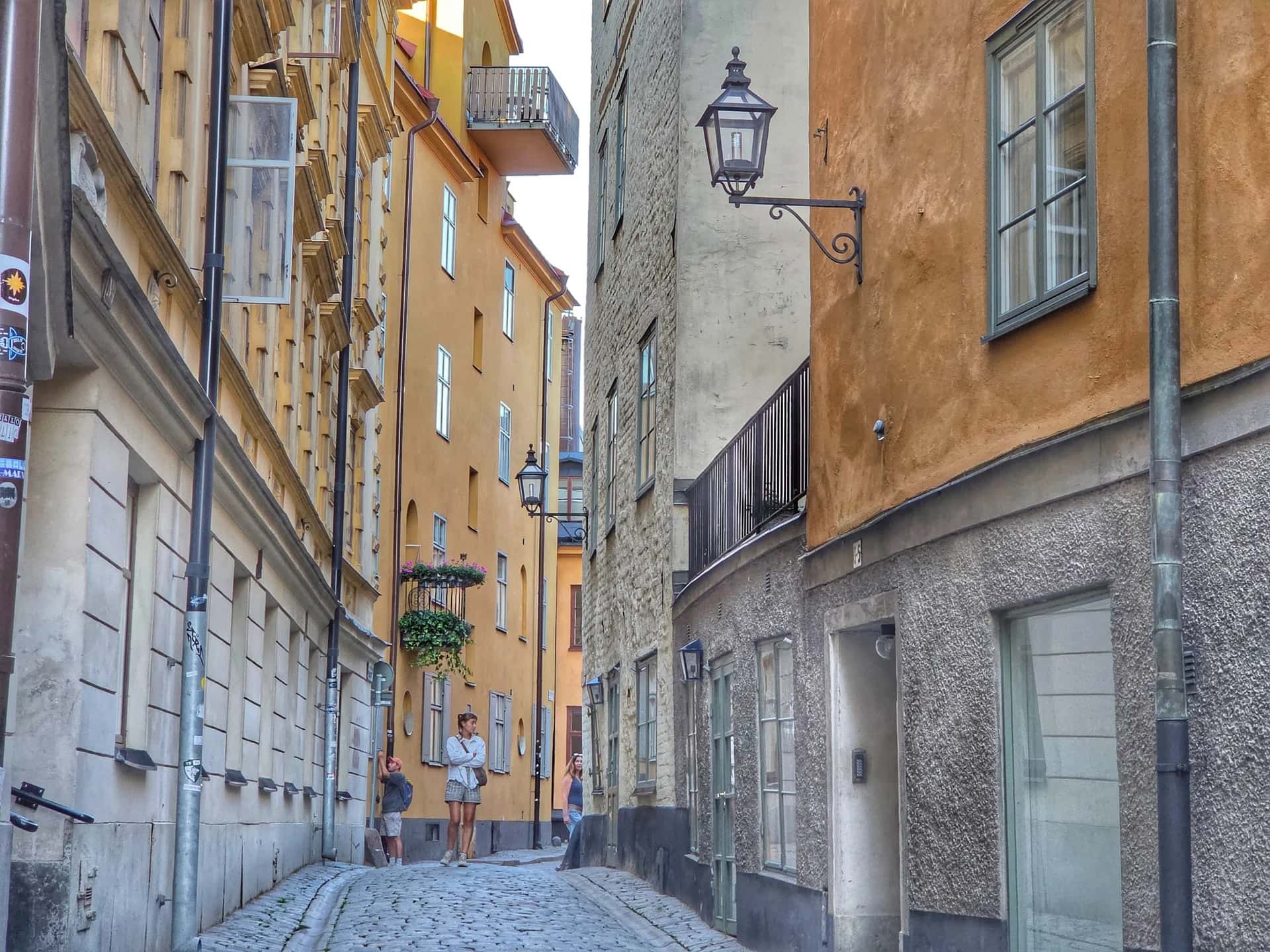Smal kullerstensgata kantad av gamla byggnader i gult och orange. Flera personer promenerar. Arkitektur Gamla stan