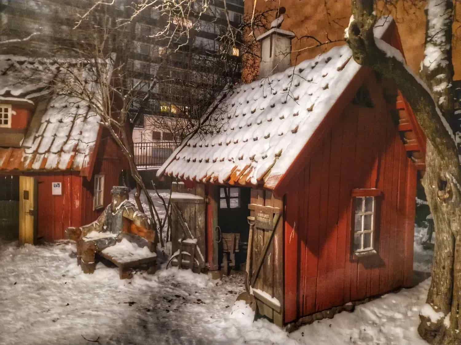 Två röda stugor med snö på taken. Snö på marken. Träd och hus i bakgrunden