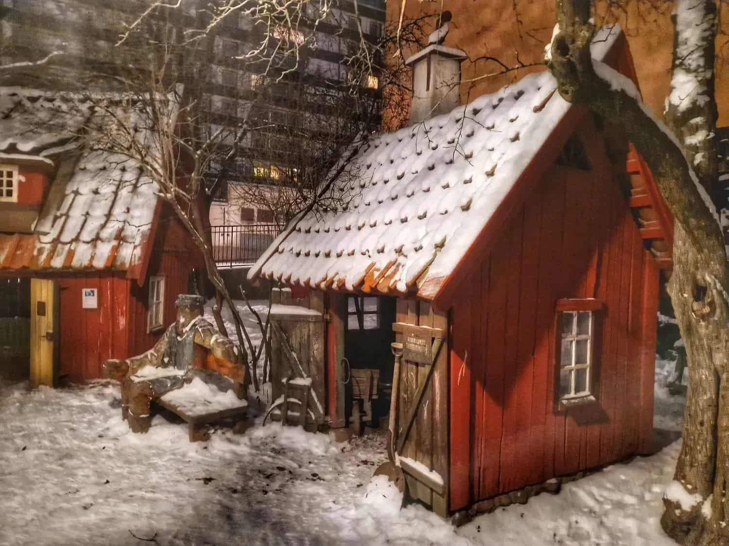 Two red cottages with snow on the roofs. Snow on the ground. Trees and houses in the background