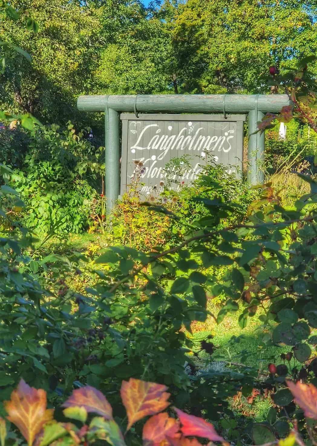Wooden sign with the text "Långholmen's florist garden" among greenery