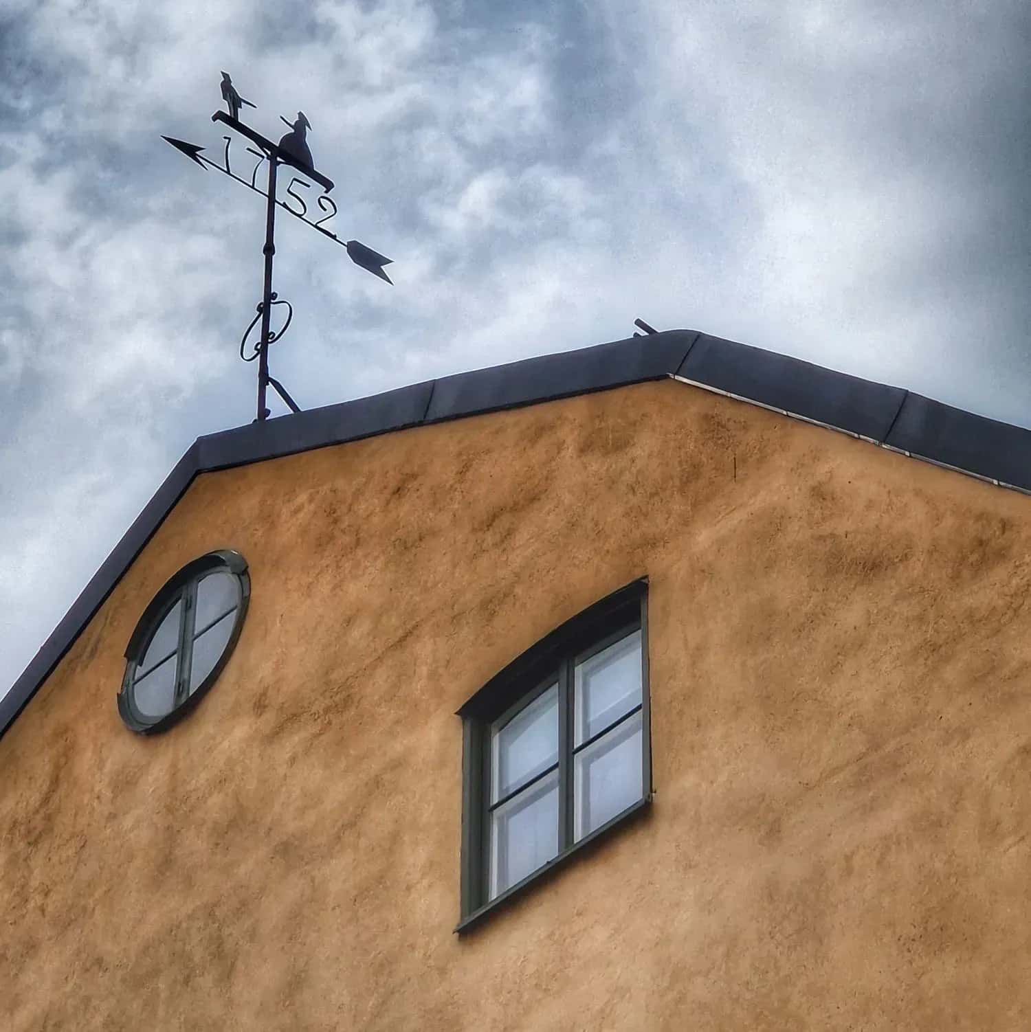 Gavel med två fönster, vindflöjel med årtalet 1752 och två siluetter. Molnig himmel