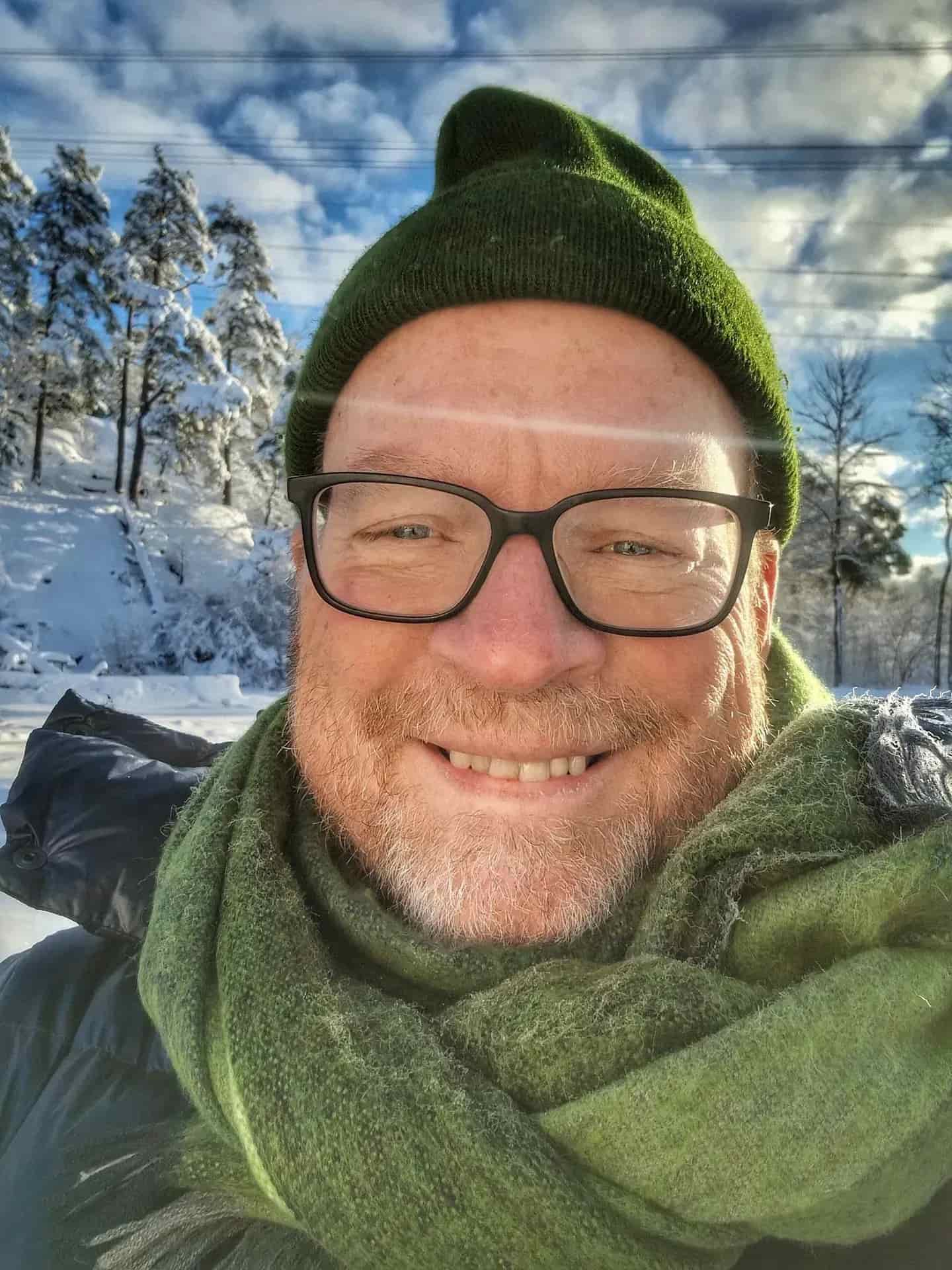 Leende man med glasögon, grön mössa och halsduk utomhus. Snö och träd i bakgrunden
