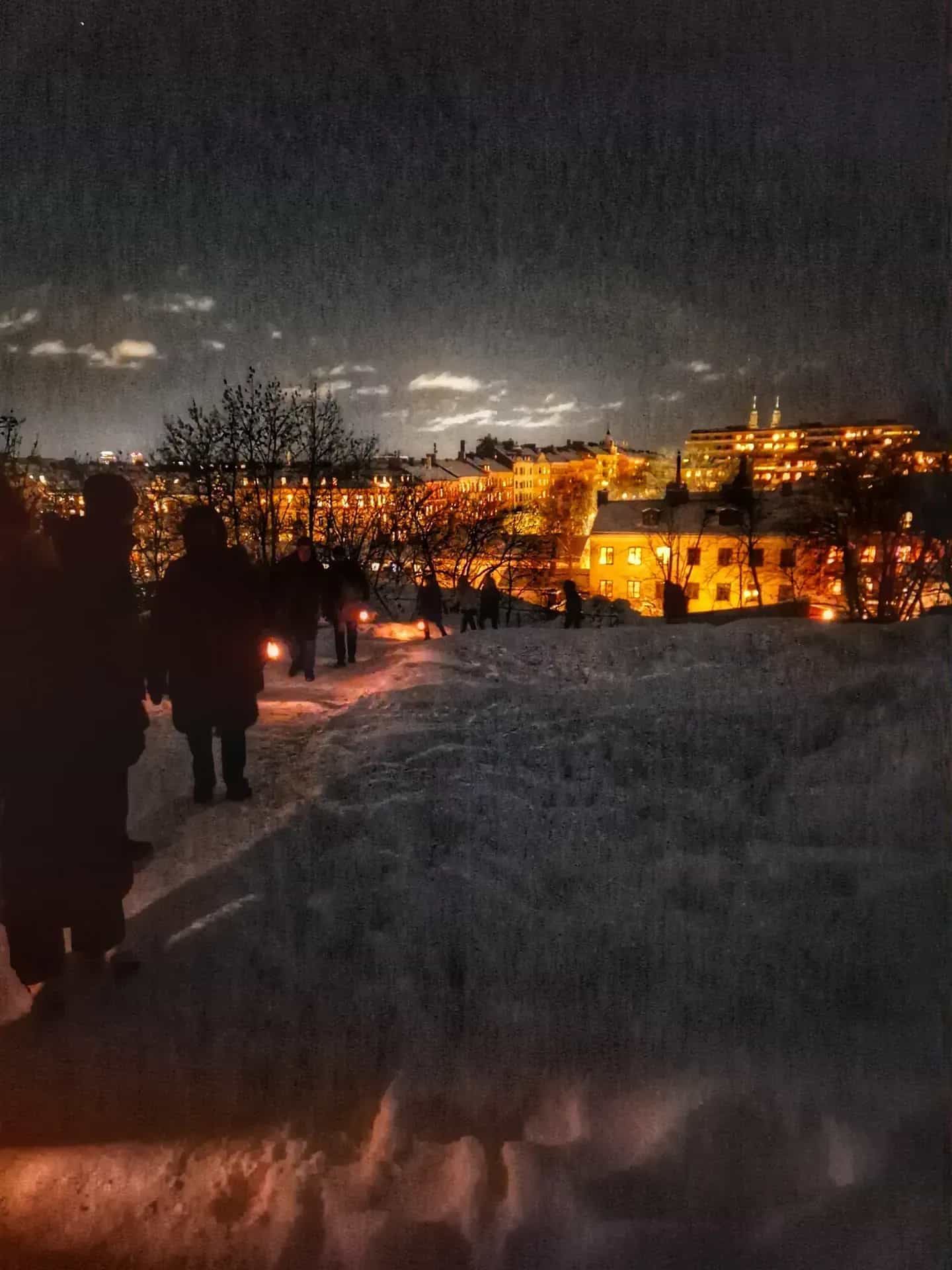 Mörkt vinterlandskap. Silhuetter av människor med lyktor i snön. Upplysta byggnader i bakgrunden