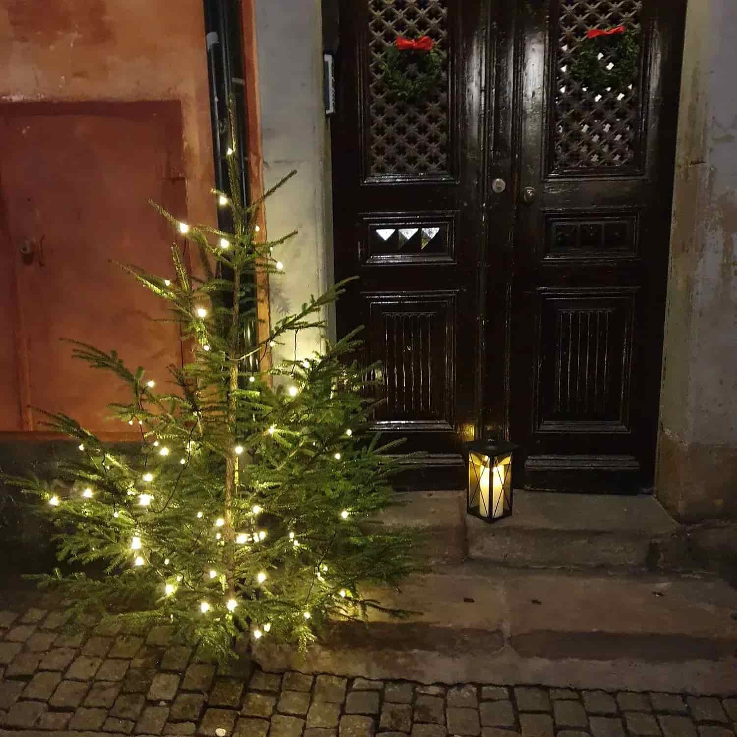 Julgran med ljusslinga, lykta och kransar vid en dörr