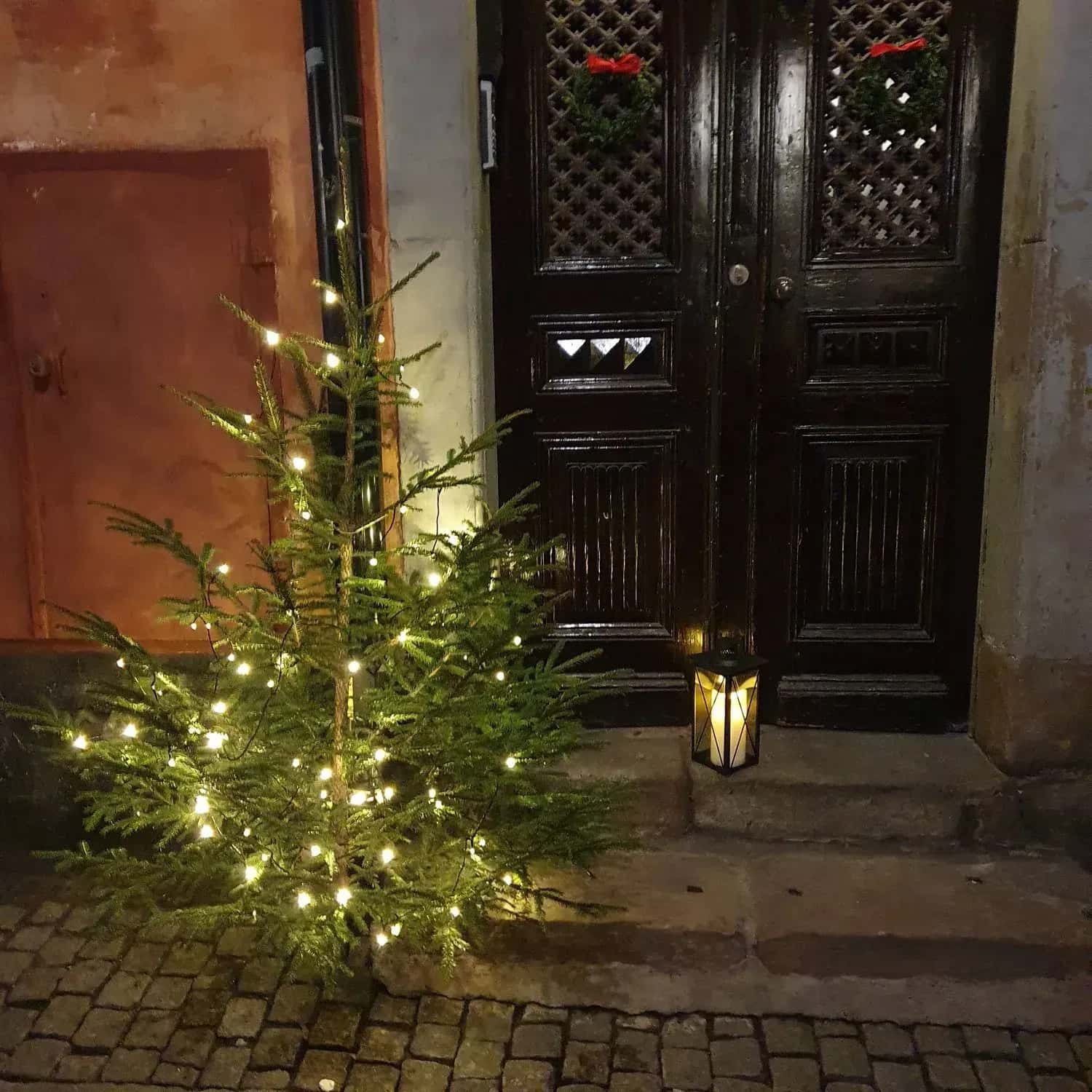 Julgran med ljusslinga, lykta och kransar vid en dörr
