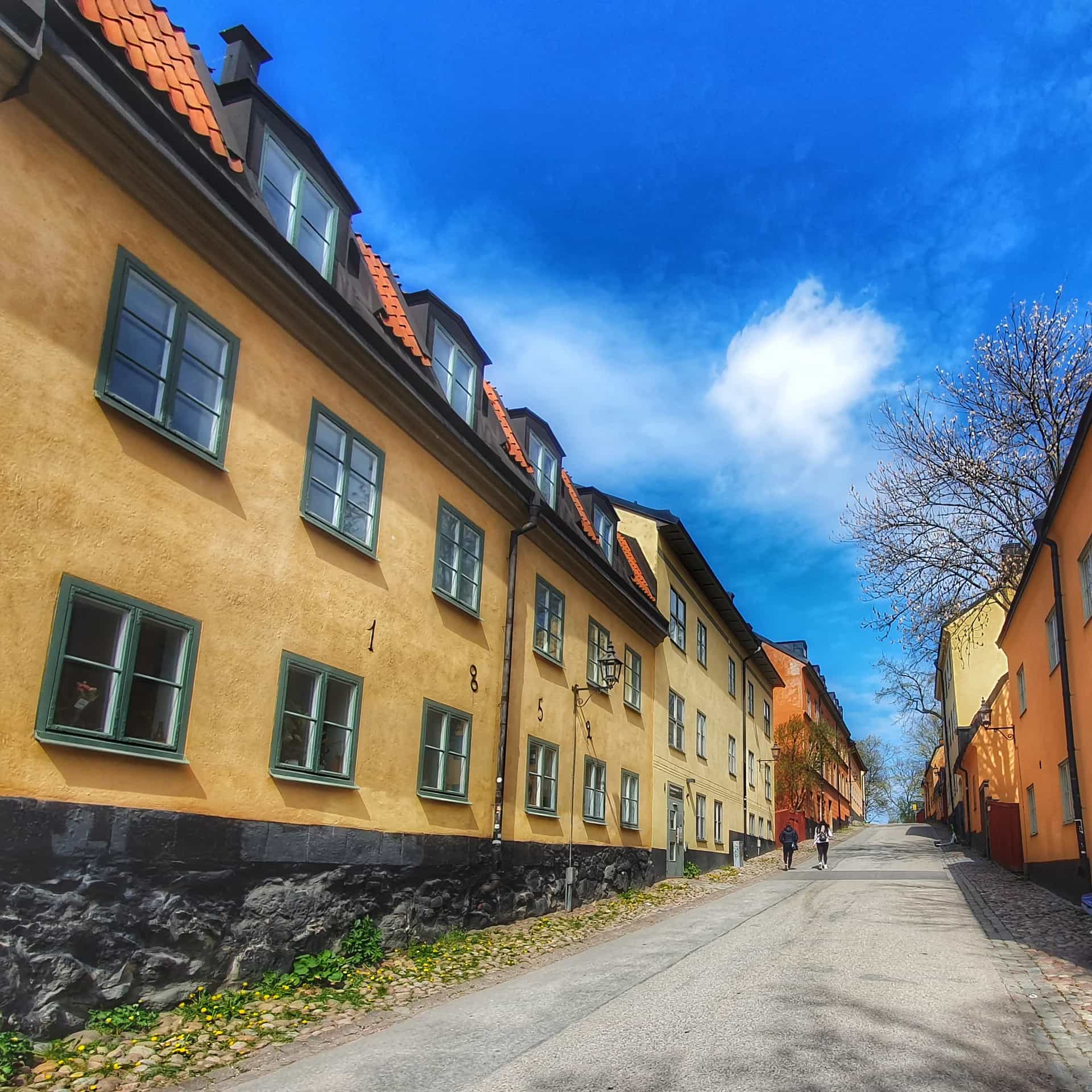 Smal gata med gula hus och tegeltak på Mariaberget, Södermalm, under blå himmel. Två personer går uppför backen