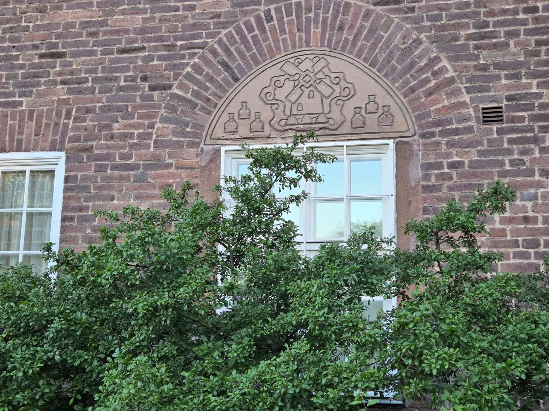 Detalj från stadsvandring Stockholm: Tegelvägg med fönster och en relief av väderkvarn med flaskor. Gröna buskar