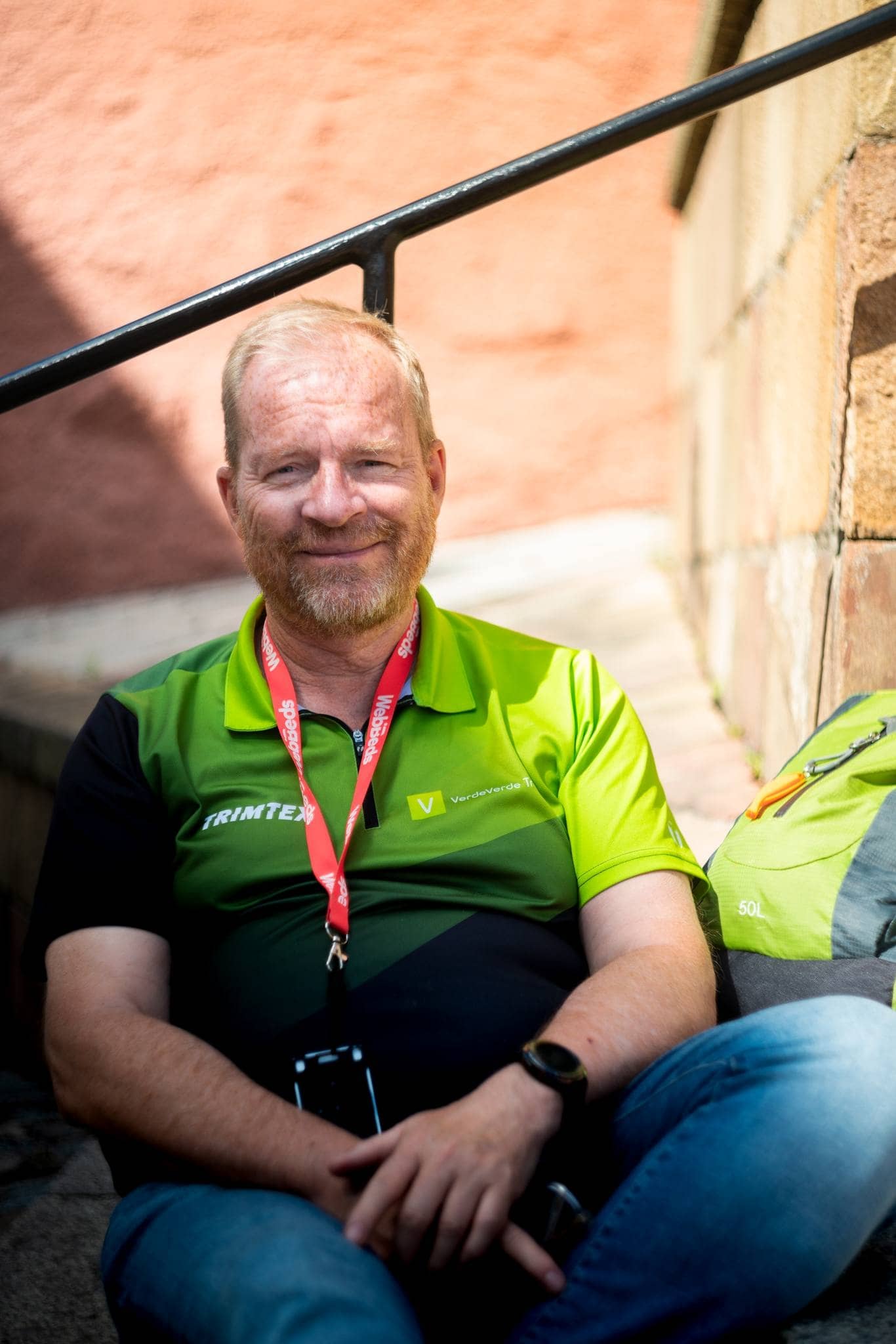 Man in green sweater with red band around his neck sitting outdoors. Backpack next to him