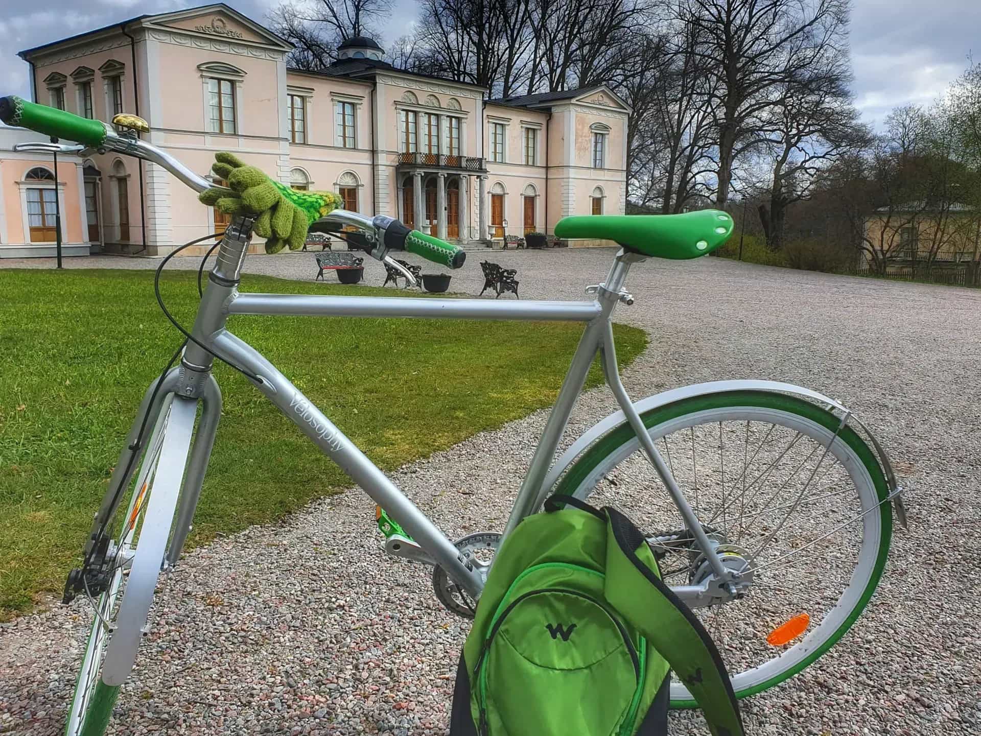 Cykeltur Stockholm. Cykel med grön sadel och handskar. Byggnad i bakgrunden. Grusväg