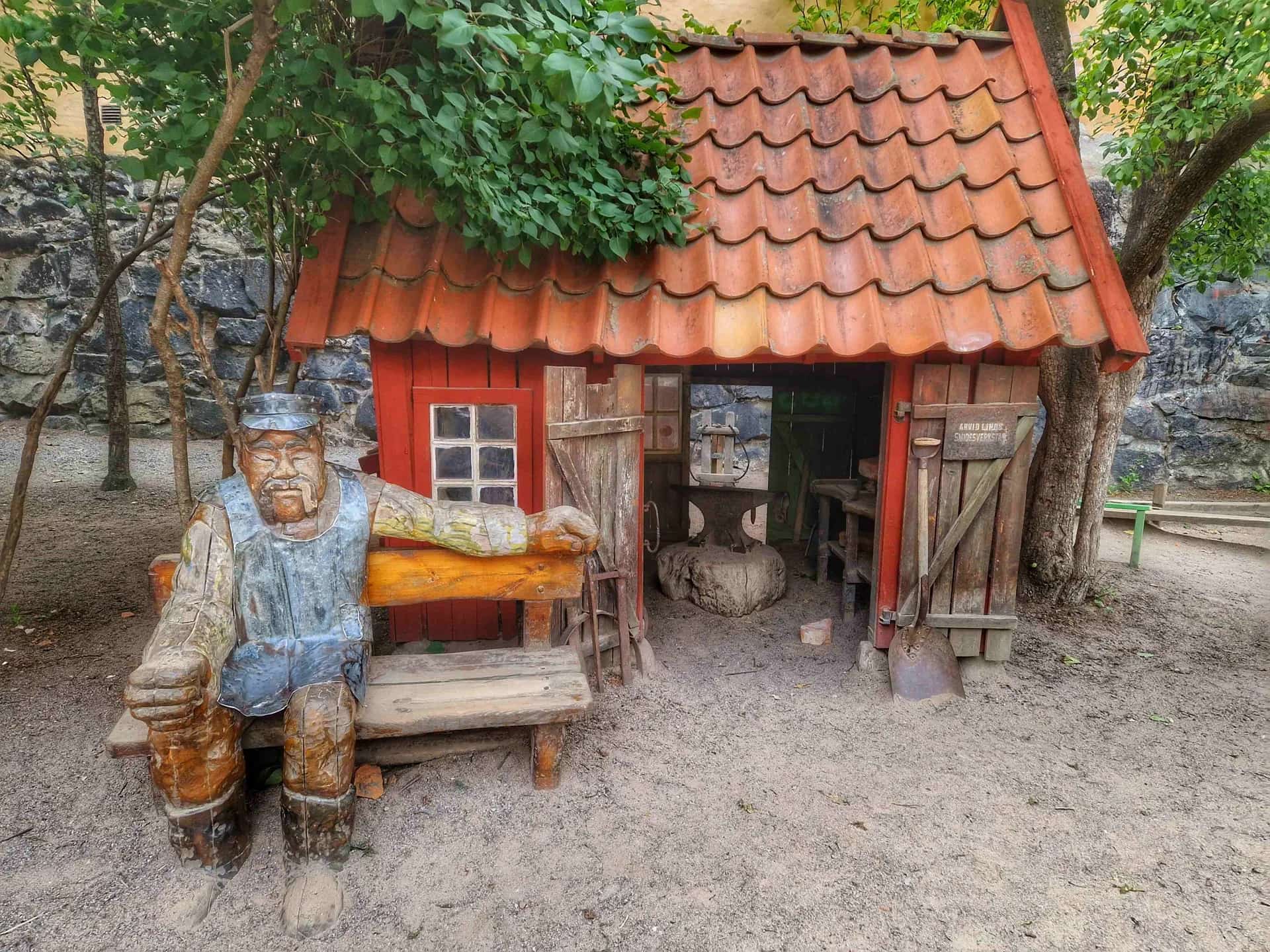 Wooden sculpture of a man on a bench, next to a smithy with a red tile roof