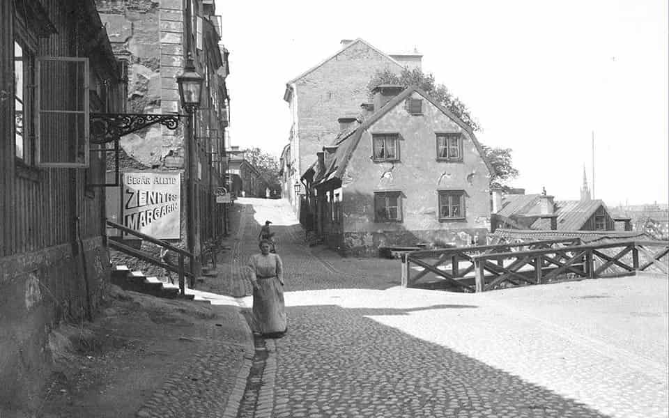 Street with cobblestones, houses and a woman in a long dress. Sign for Zenith Margarin