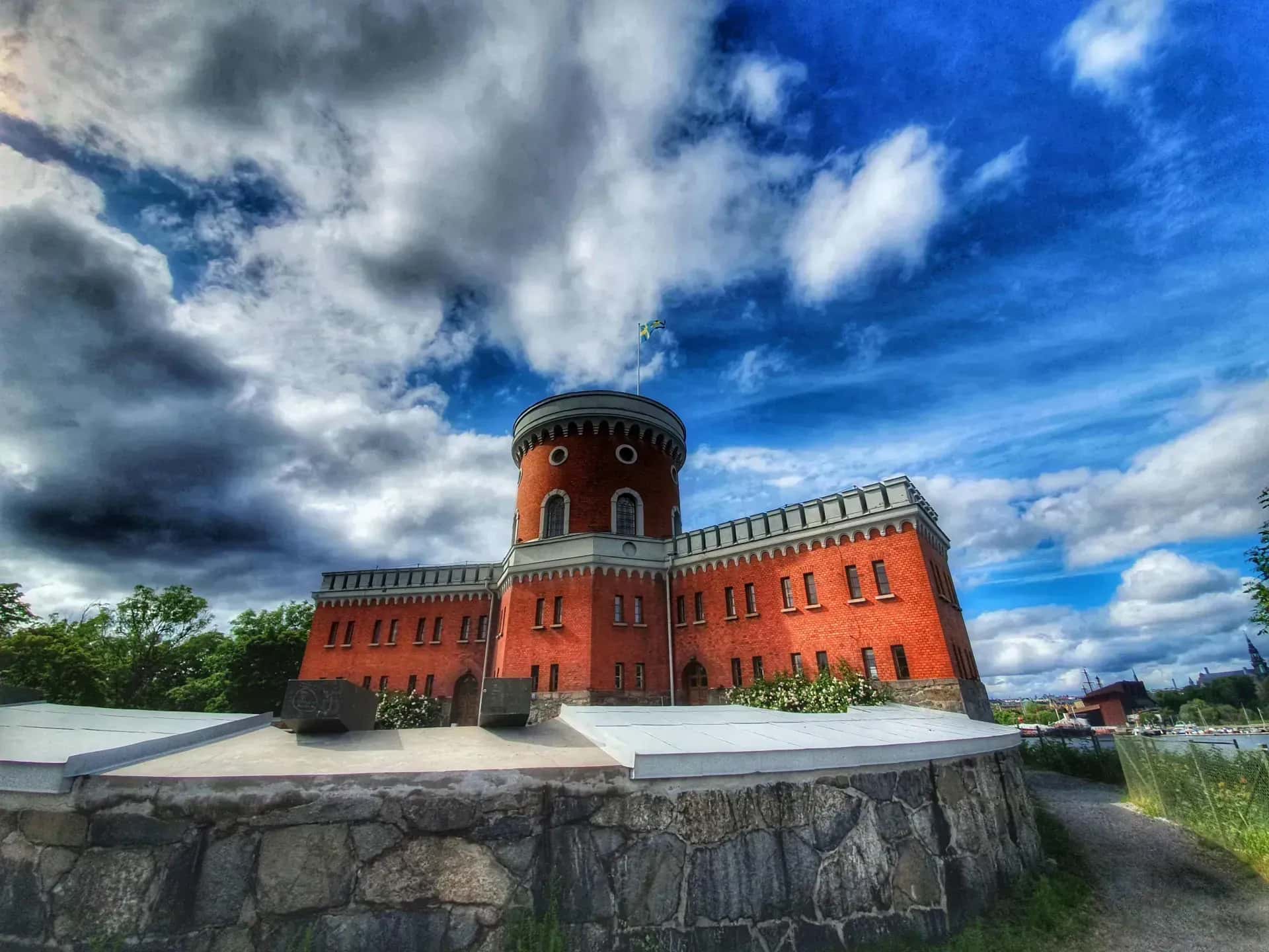 Kastellholmen, Stockholm. Röd tegelbyggnad med torn och svensk flagga mot himmel med moln