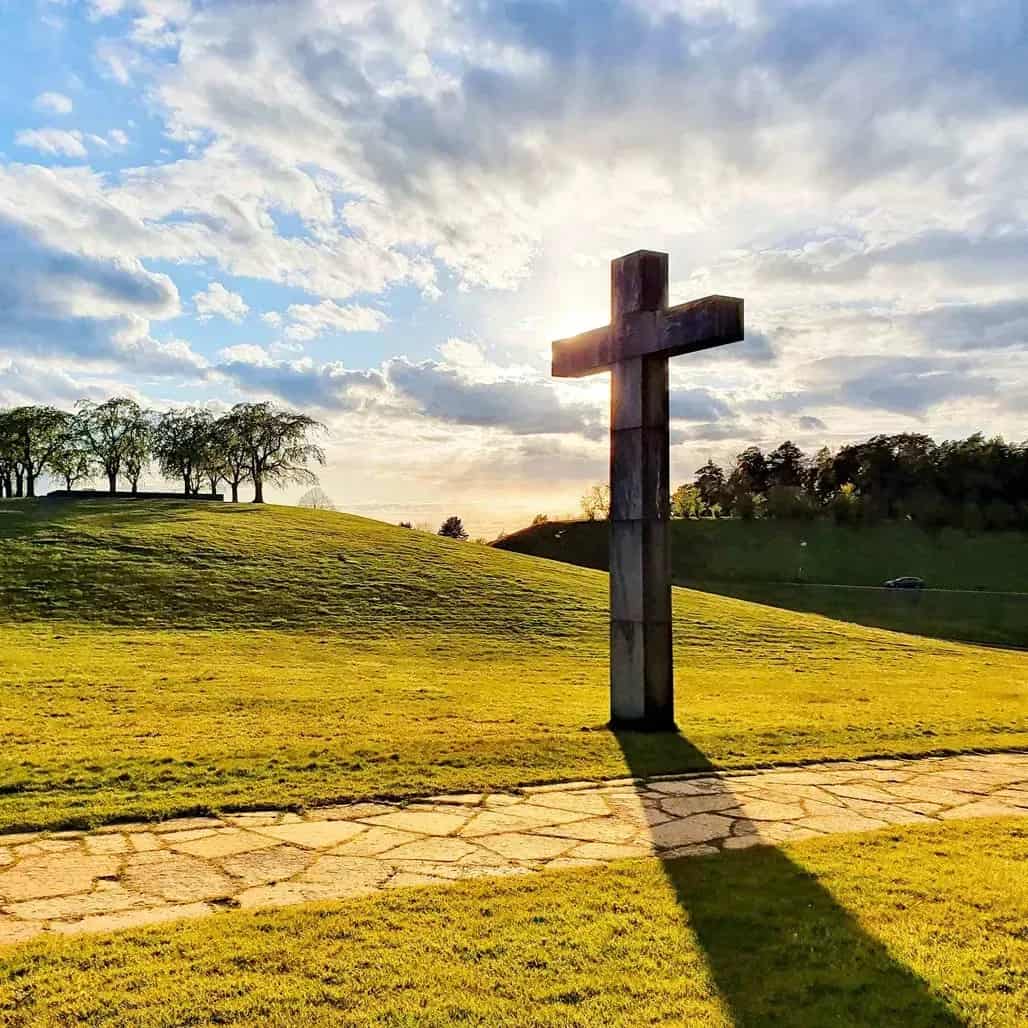 Large concrete cross on a grassy hill, stone-paved walkway. Sun and clouds in the sky