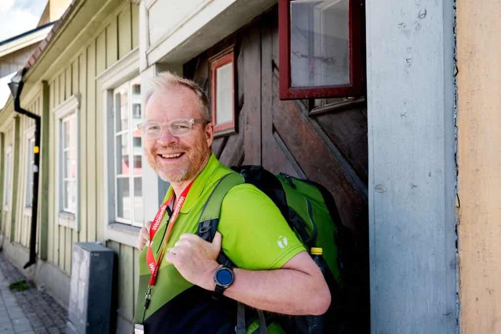 Leende man med ryggsäck och grön tröja. Han har glasögon och ett rött band runt halsen
