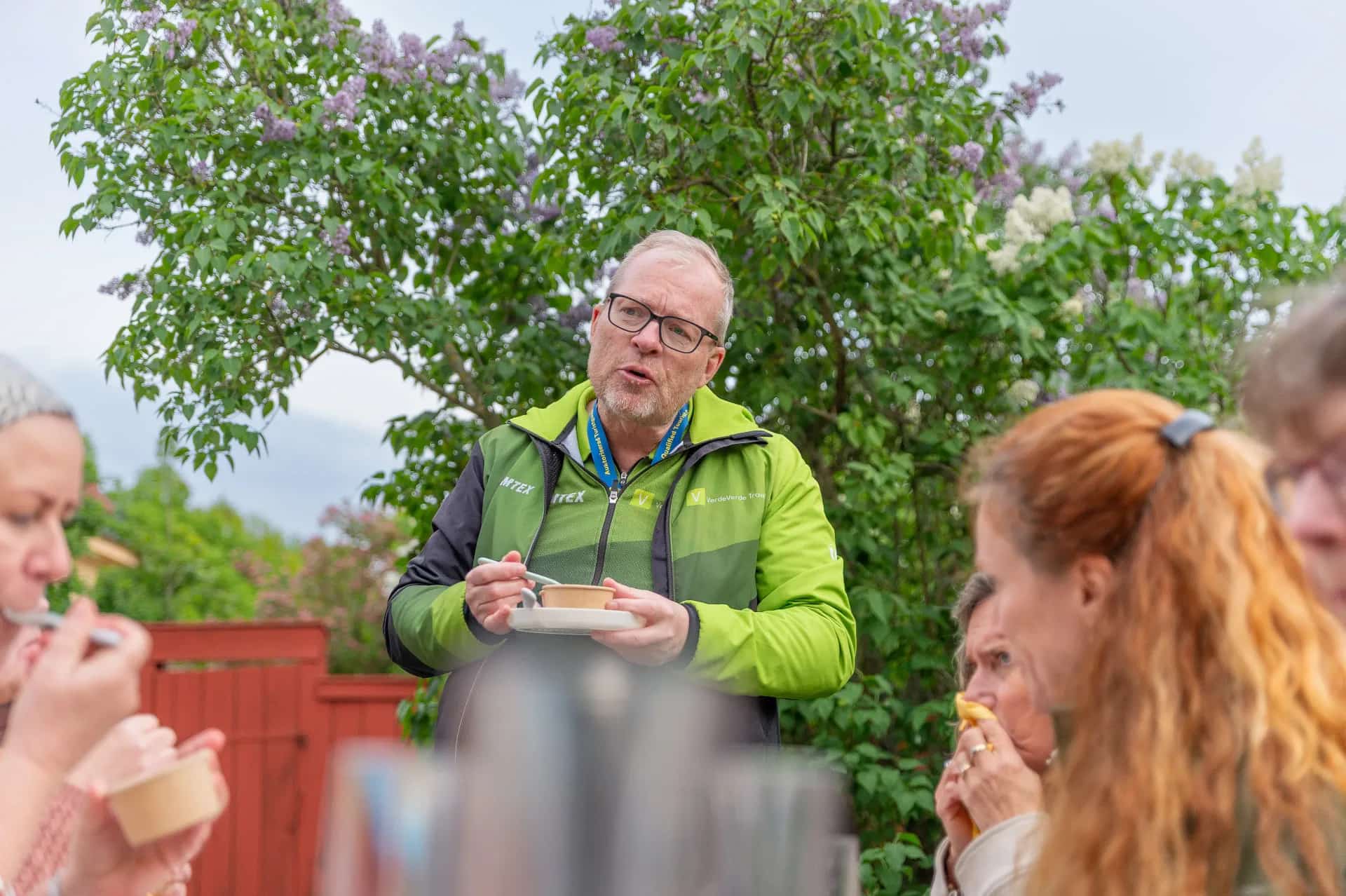 Man holds plate with bowl and spoon outdoors, others eat. Lilacs in the background