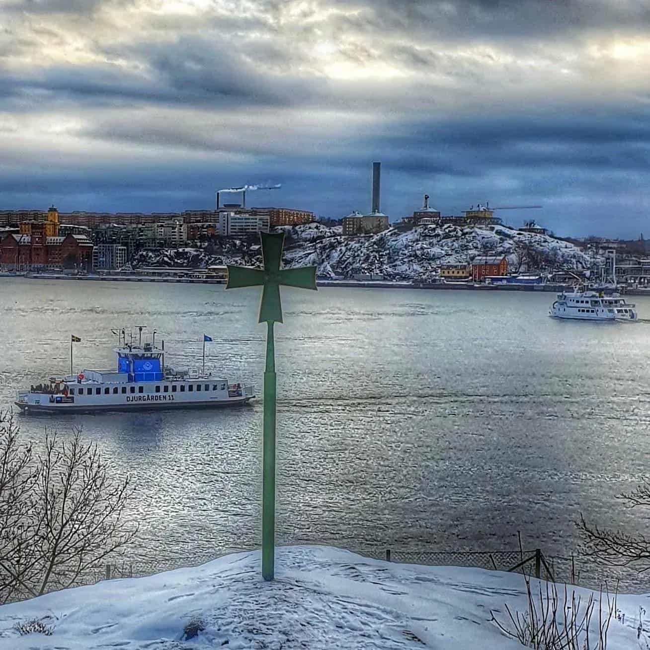 Winter view of water with two boats, city in the background and a green cross in the foreground