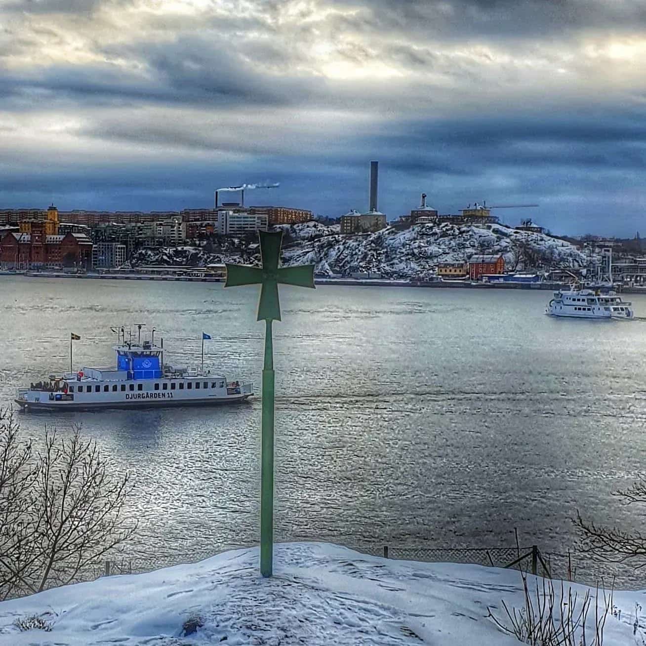 Winter view over water with two boats, city in the background and a green cross in the foreground