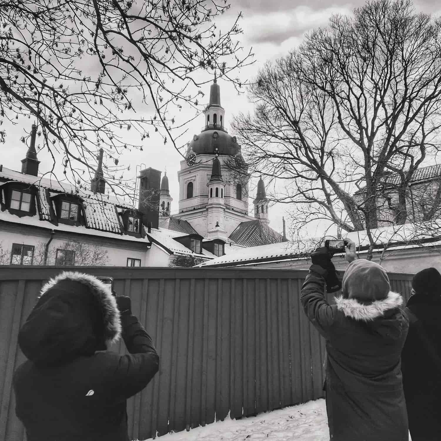Tre personer fotograferar en kyrka med torn bakom ett staket i svartvitt