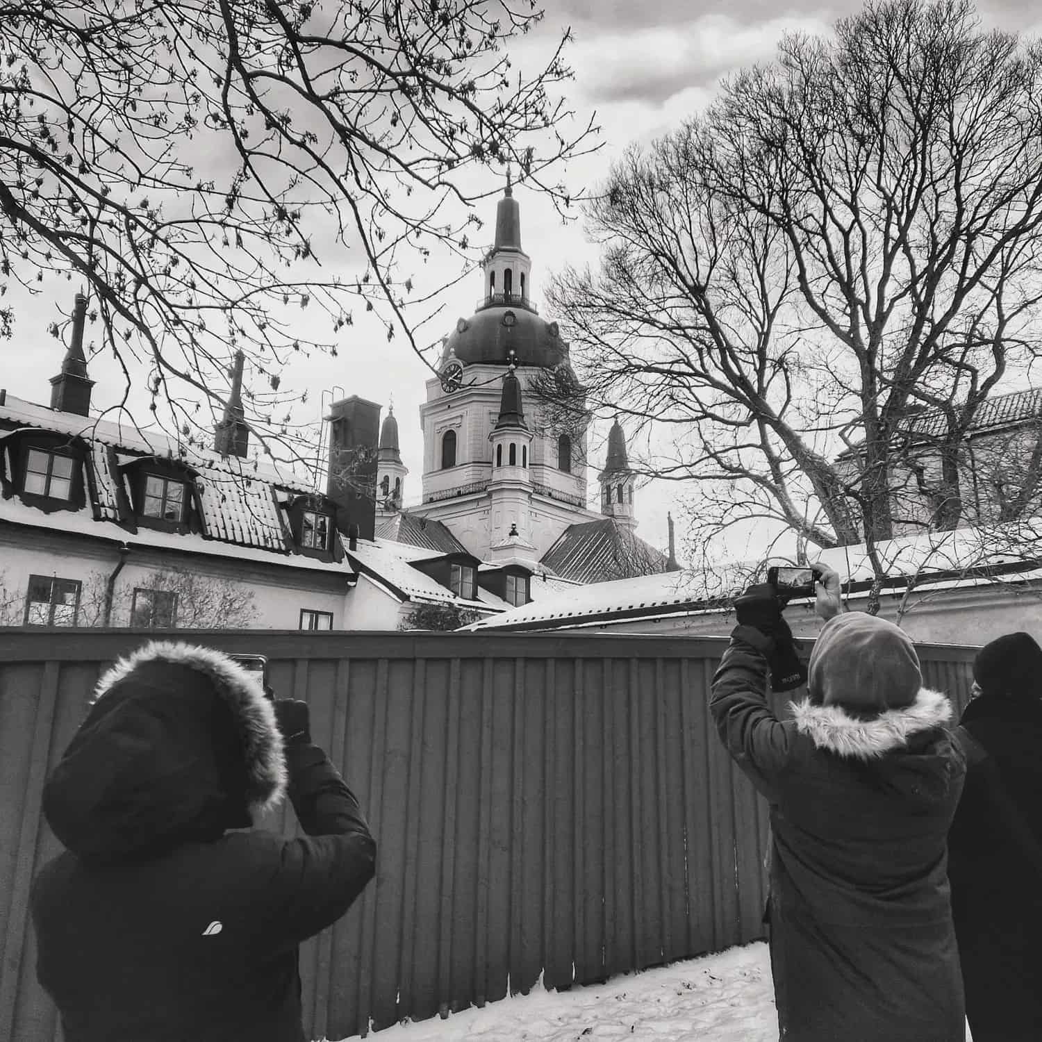 Three people photograph a church with a tower behind a fence in black and white