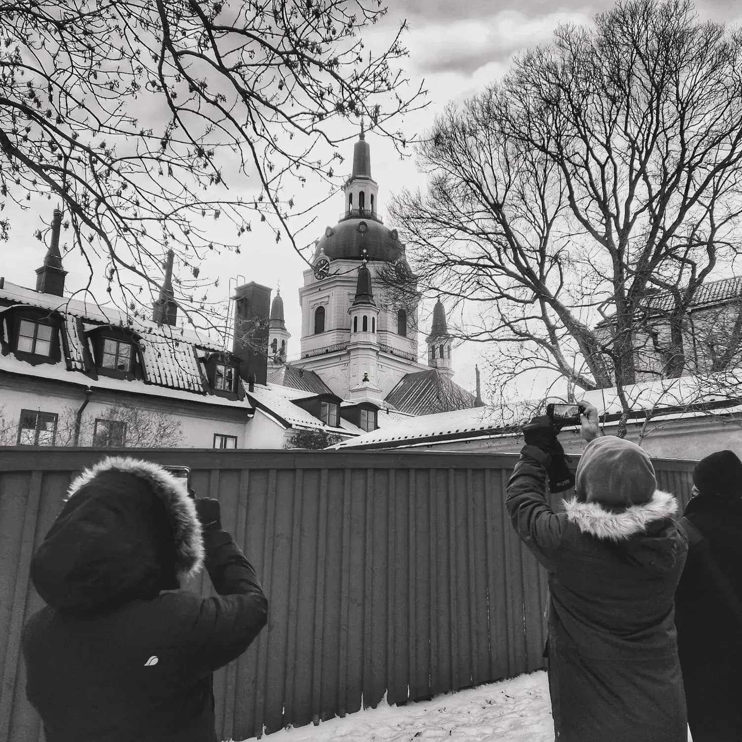 Tre personer fotograferar en kyrka med torn bakom ett staket i svartvitt
