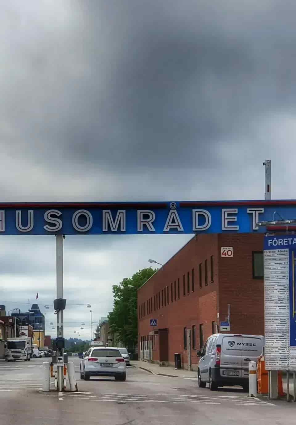 Blå skylt "HUSOMRÅDET" över väg med bilar och rött tegelhus