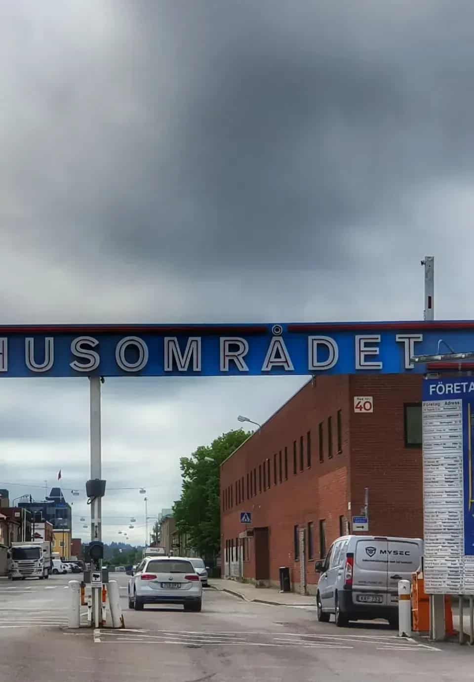 Blå skylt "HUSOMRÅDET" över väg med bilar och rött tegelhus