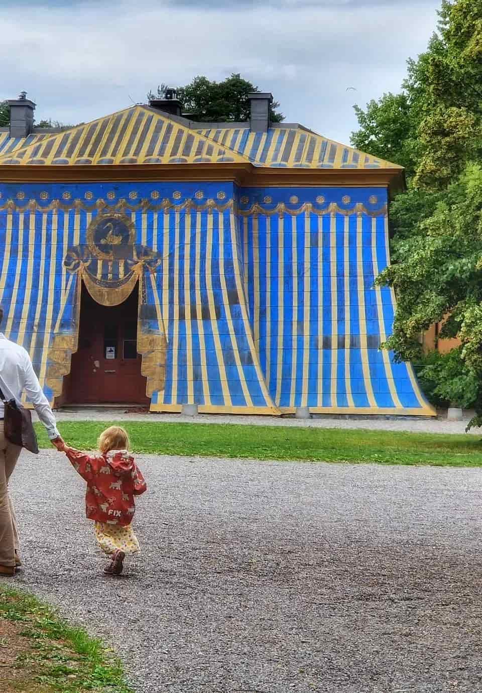 Man och barn hand i hand framför byggnad målad som tält i blått och gult