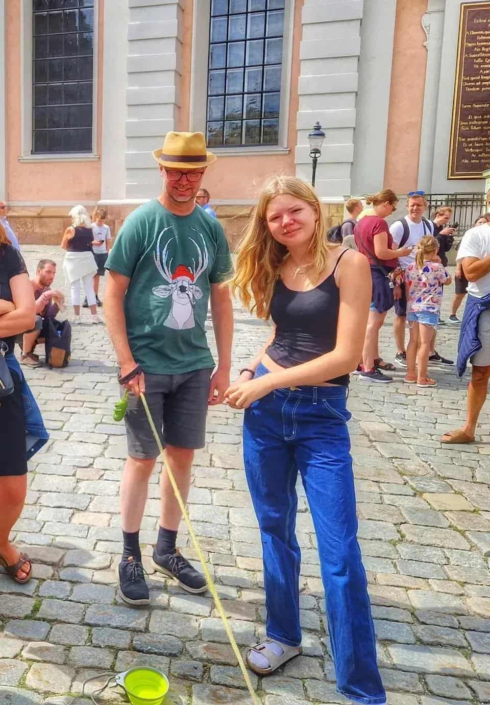 Woman and man on cobblestones. The man is wearing a hat and a t-shirt with a reindeer. Leashed green bowl. Several people in the background