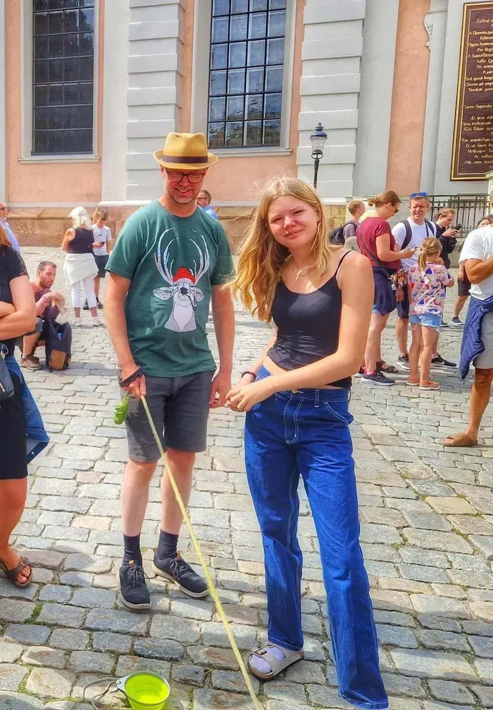 Kvinna och man på kullersten. Mannen har hatt och t-shirt med ren. Kopplad grön skål. Flera personer i bakgrunden