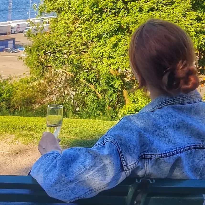 Kvinna med jeansjacka sitter på bänk med champagneglas, blickar ut över hamn