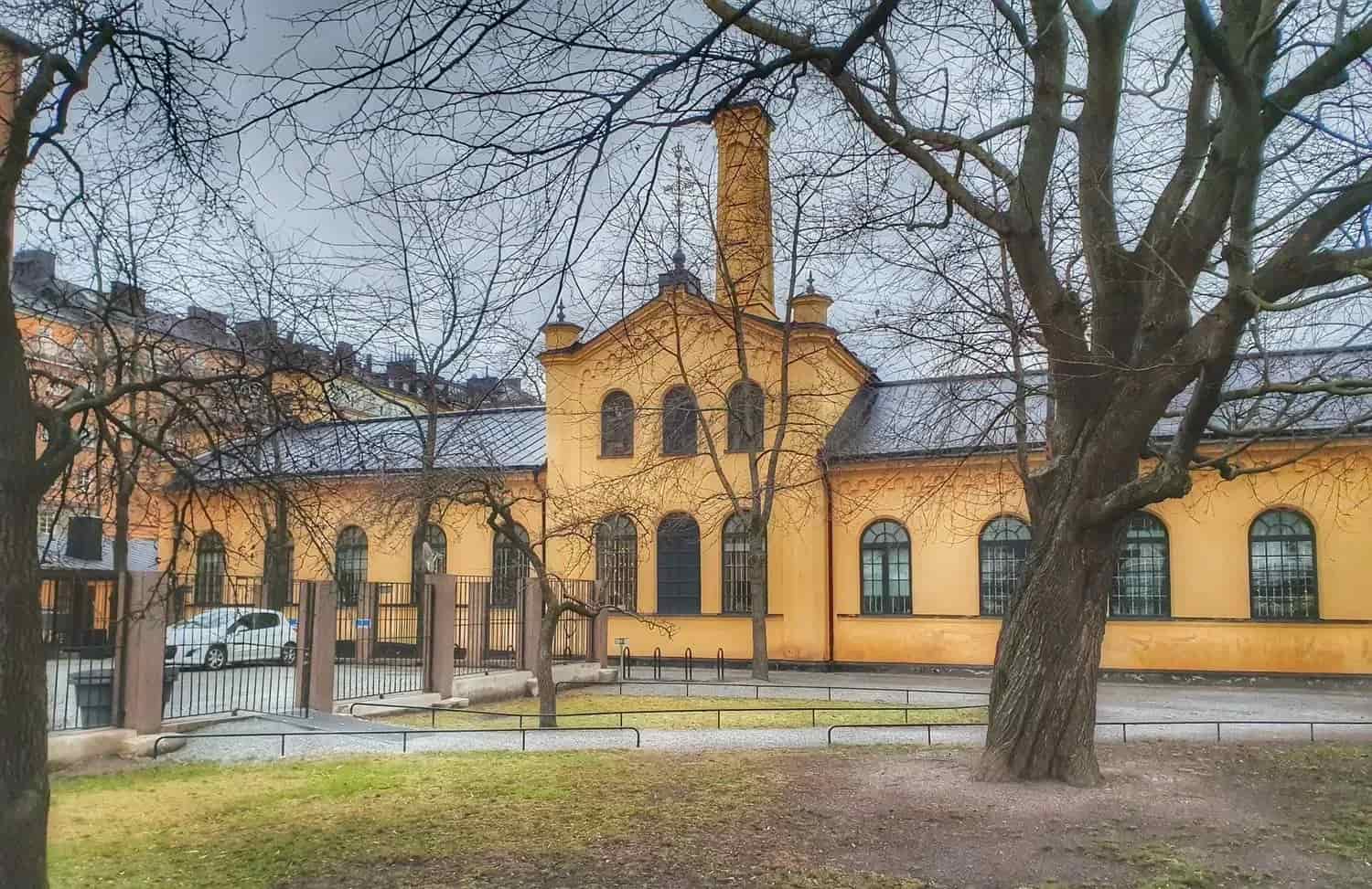 Gul byggnad med tegeltak bakom träd i park. Molnig himmel