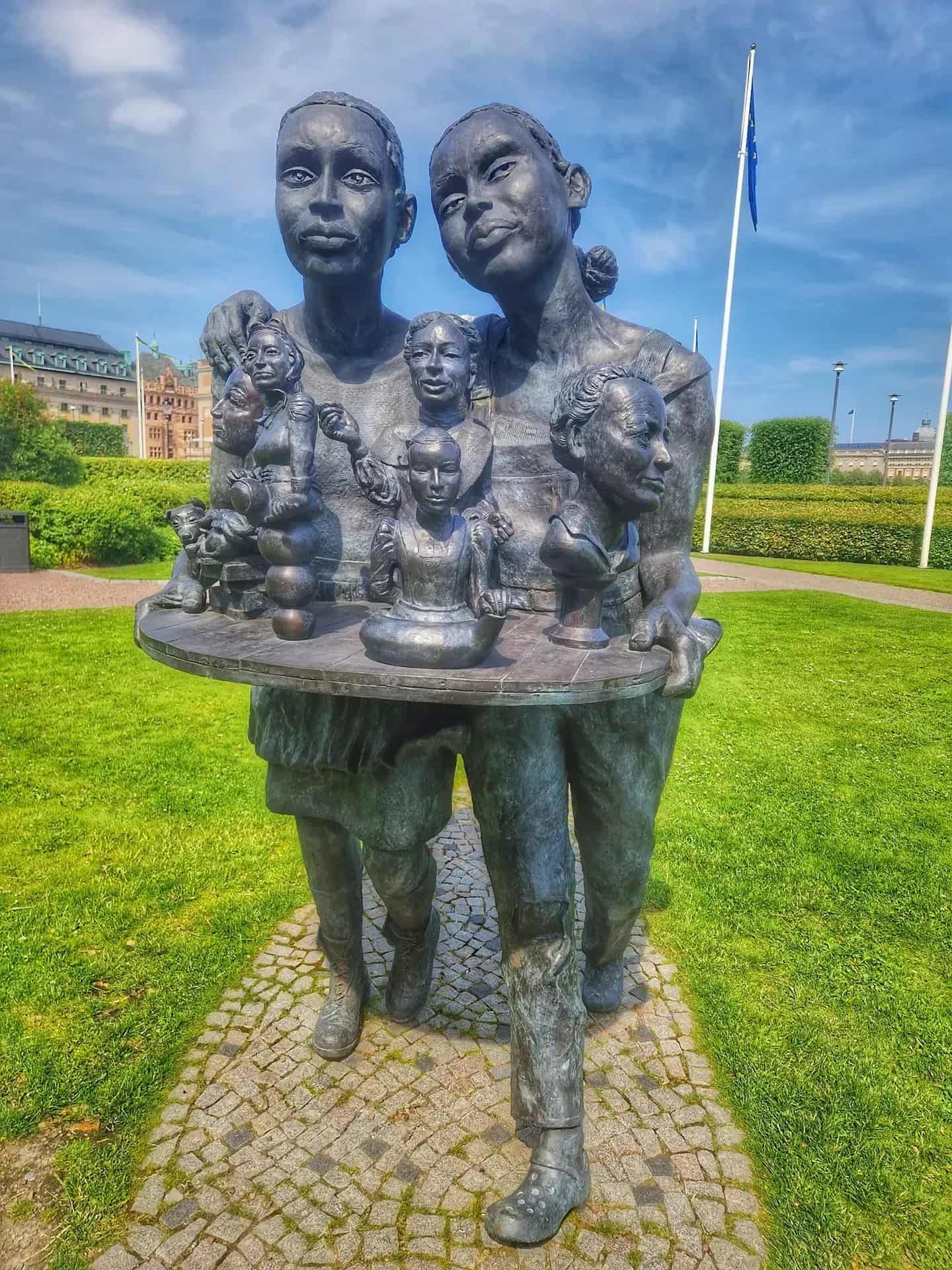 Bronze statue of two girls carrying a round table with busts. Grass and buildings in the background