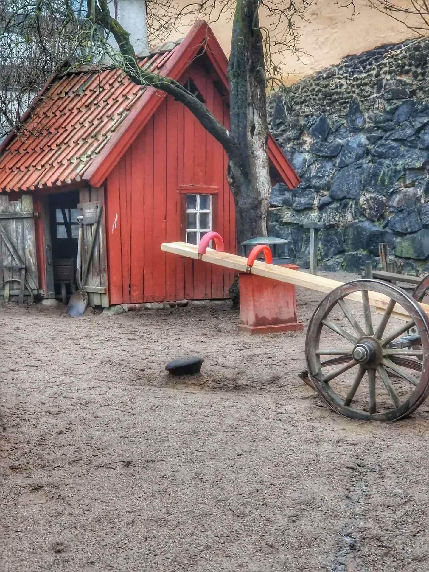 Röd liten stuga med tegeltak, gungbräda och vagnshjul på grus. Stenmur i bakgrunden