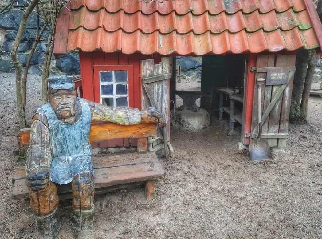 Träskulptur vid röd stuga med tegeltak. Inuti stugan syns ett städ