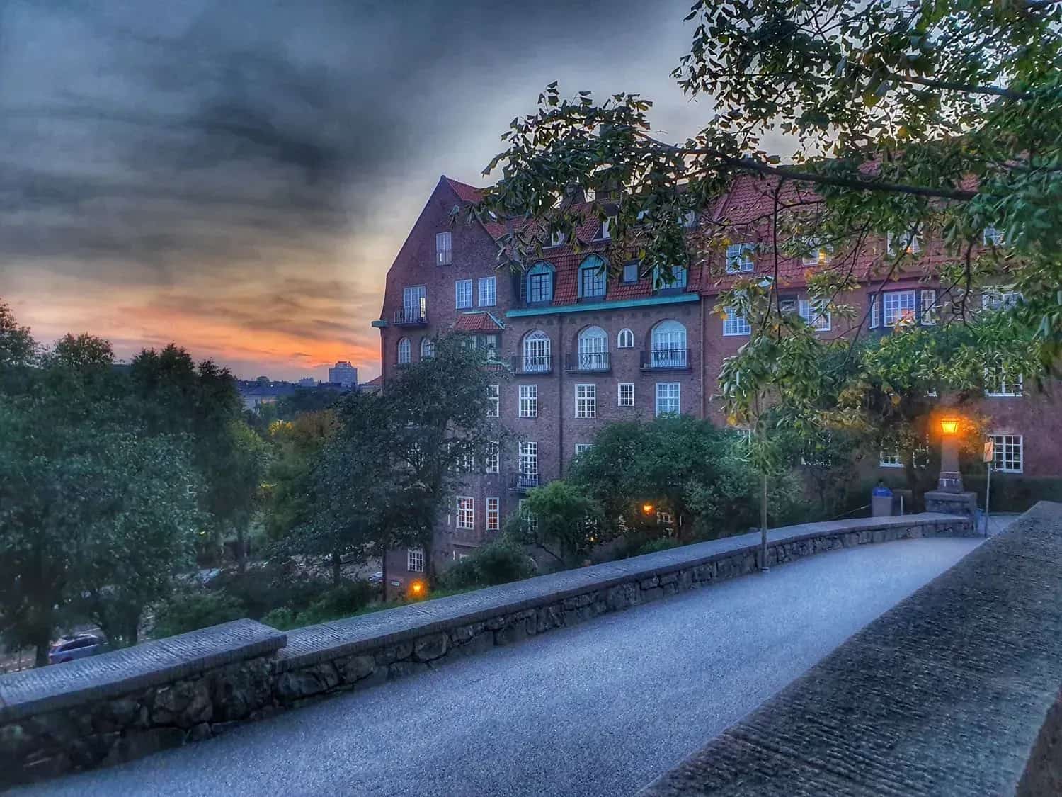 Paved walkway with stone obstacles, brick building and trees under a dramatic sky