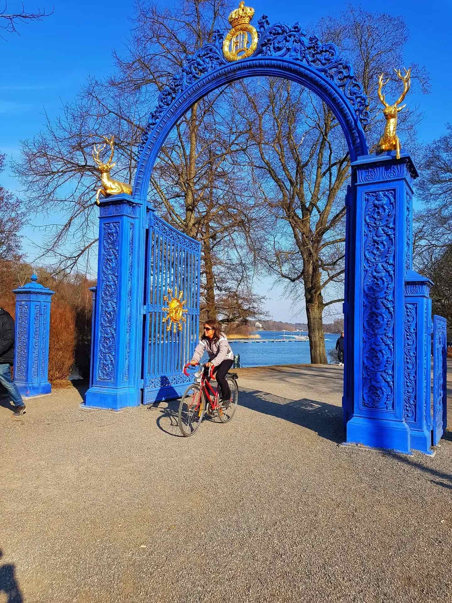 Blå grind med gulddekor, solsymbol och hjortar. Kvinna cyklar igenom. Vatten i bakgrunden