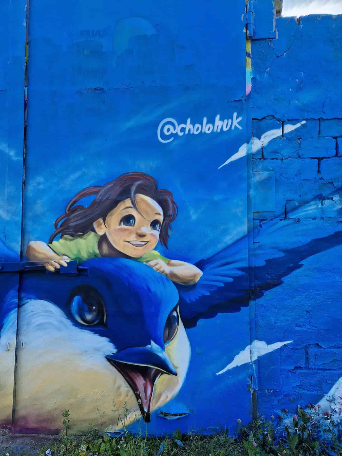 Blå väggmålning: flicka på stor blå fågel med "@cholohuk" ovanför. Moln målade i bakgrunden. Stockholm street art