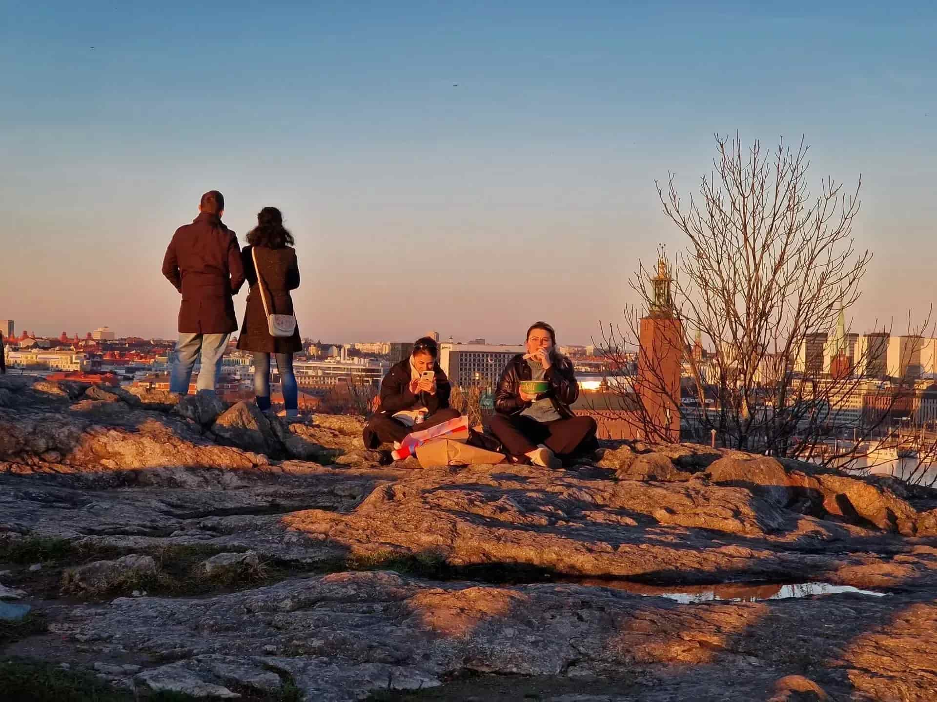 People on a mountain with a cityscape in the background. Two are sitting and eating, two are standing and looking. Sunset