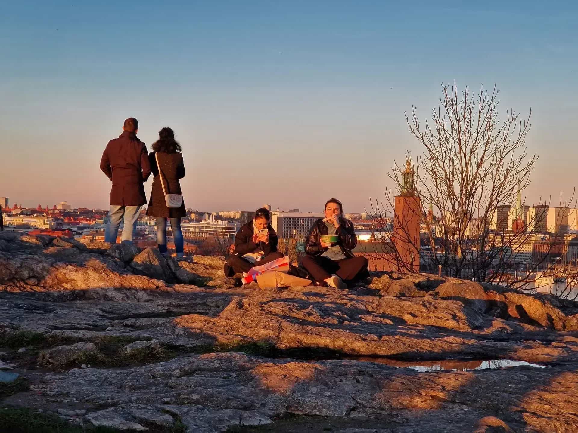 Folk på berg med stadsbild i bakgrunden. Två sitter och äter, två står och tittar. Solnedgång