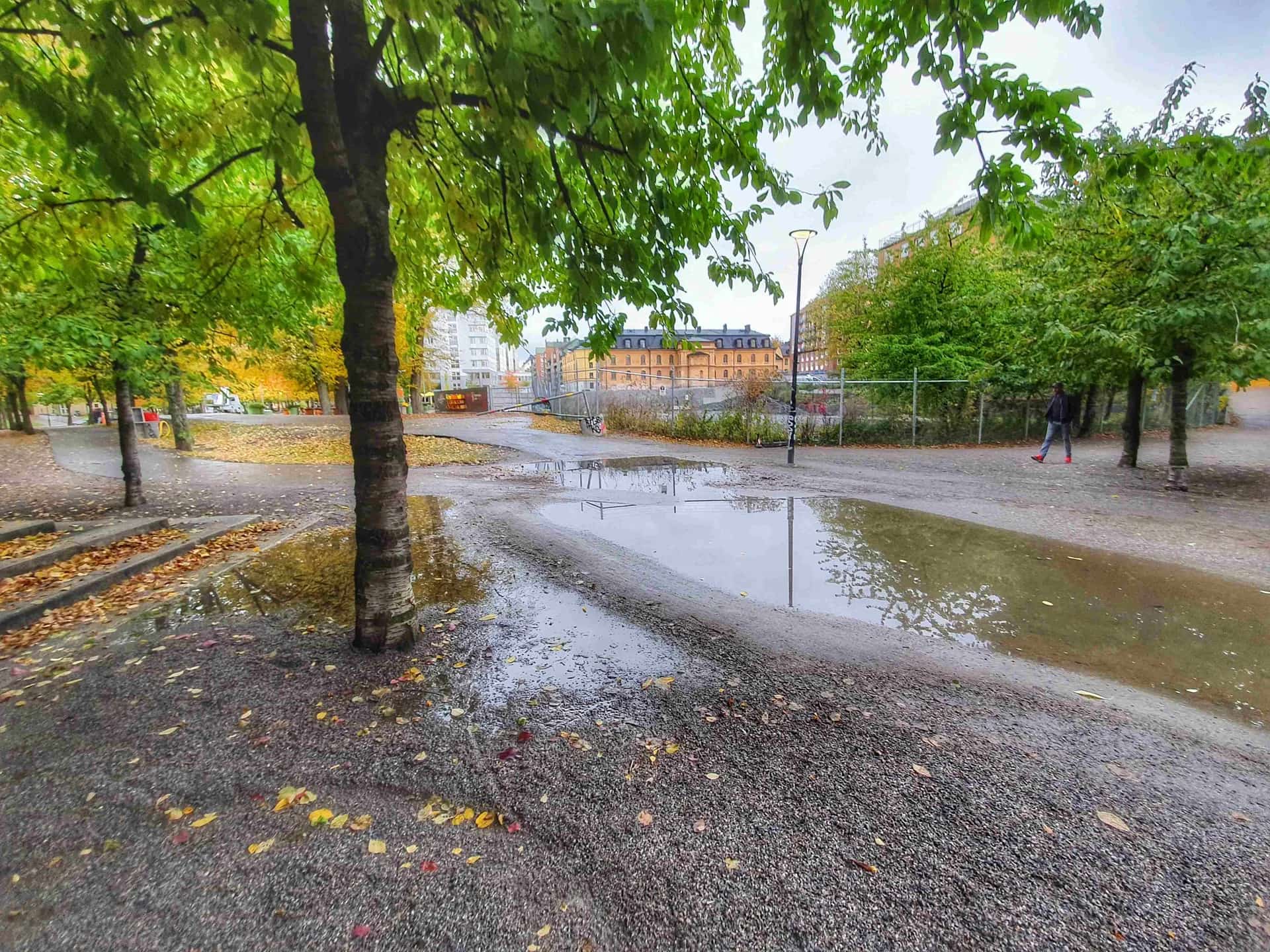 Puddle on gravel road in park. Trees, leaves and buildings can be seen in the background