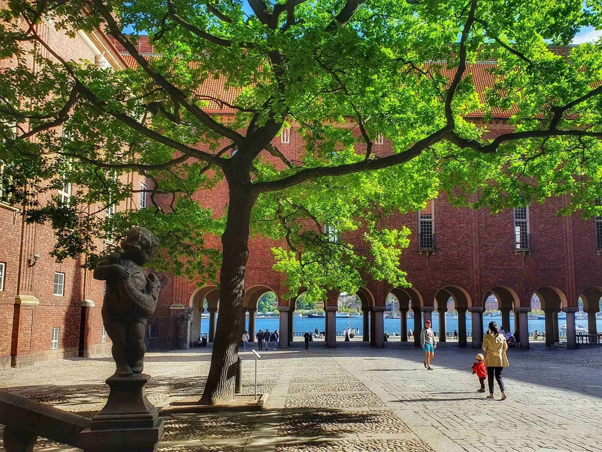 Courtyard with statue, trees and people. Brick building with arches in the background against the water