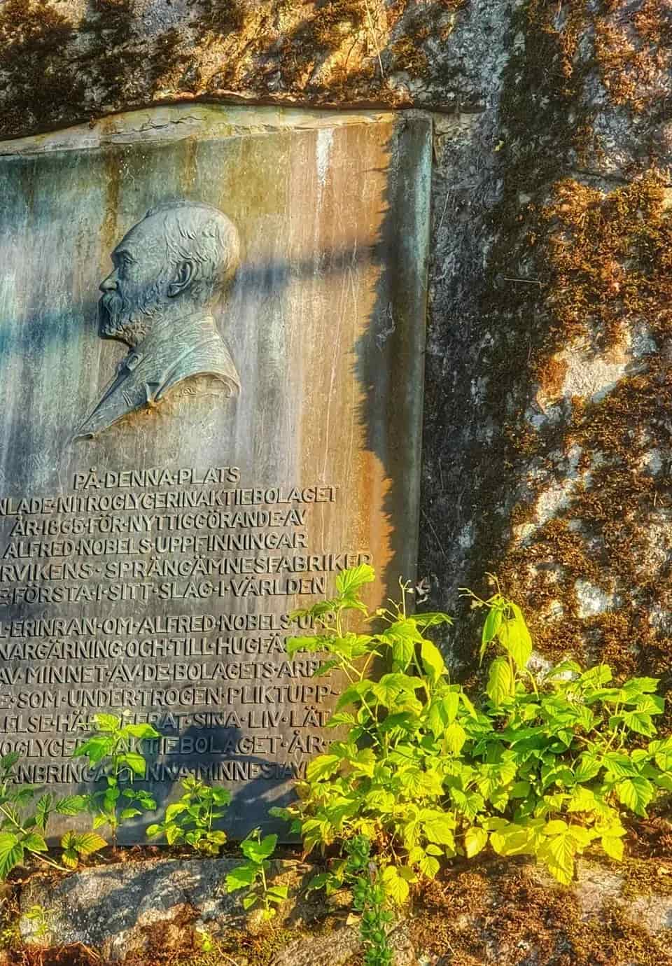 Bronsplakett med Alfred Nobel-relief och text. Växter nedanför. Mossbelagd sten bakom