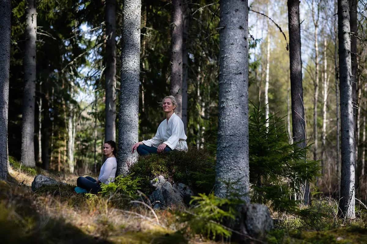 Två kvinnor sitter i lotusposition bland träden i en skog. Ekoturism Stockholm