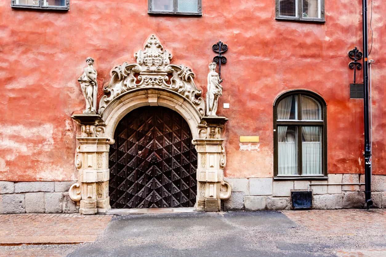 Röd husfasad med rikt ornerad stenportal, två statyer, mörk trädörr och valvformat fönster. Arkitektur Gamla stan