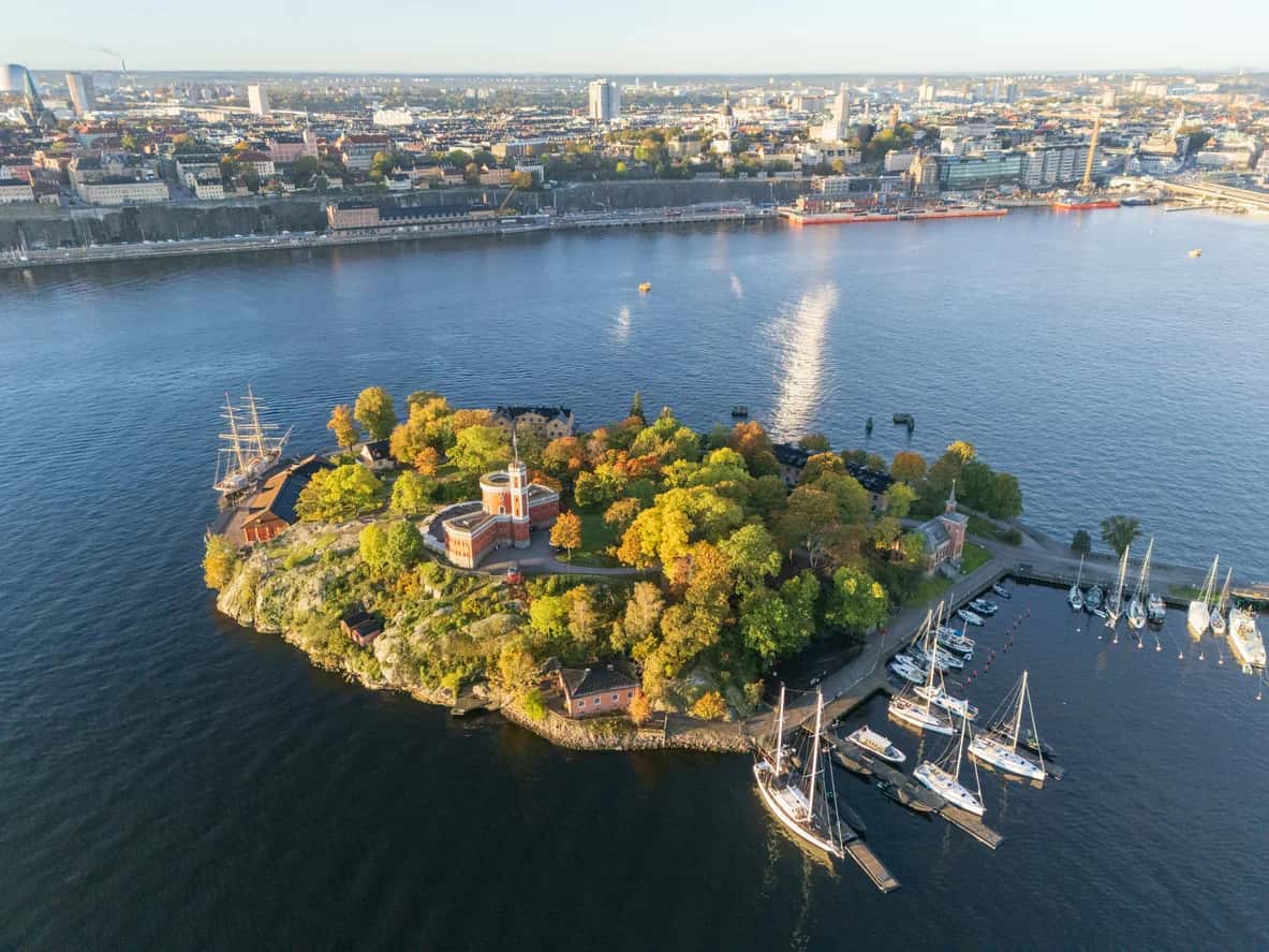 Stadsvandring Stockholm: Ö med röd borgliknande byggnad, marina, segelfartyg, höstträd. Stadssiluett i bakgrunden
