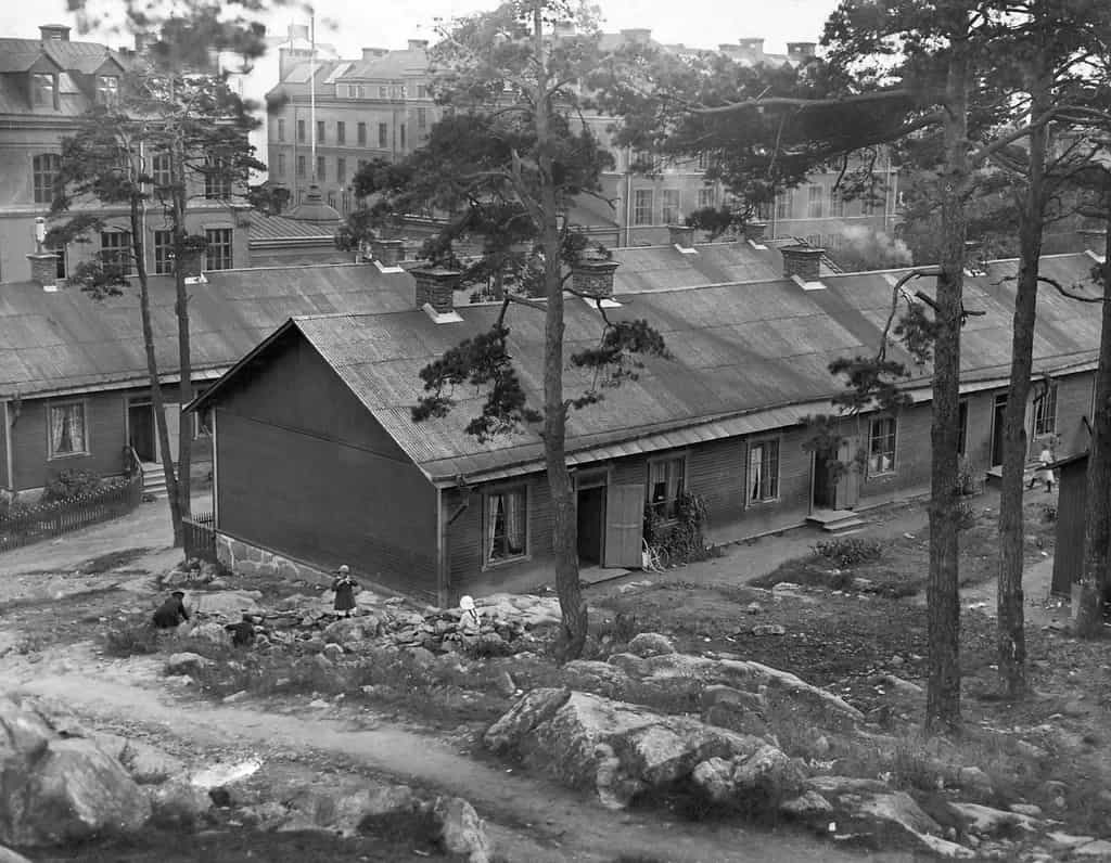 Trähus med plåttak. Flera barn leker bland stenar och träd. Större byggnader syns i bakgrunden