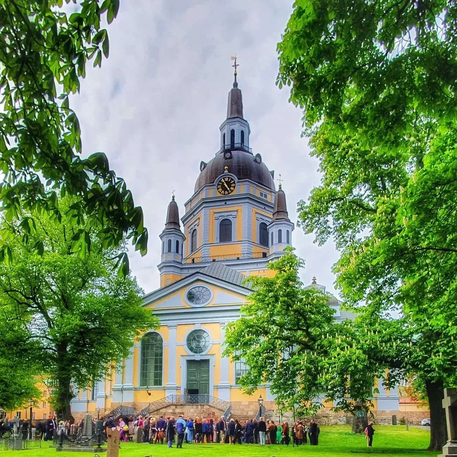 Church with tower and dome, surrounded by trees and people. Lawn and tombstones in the foreground